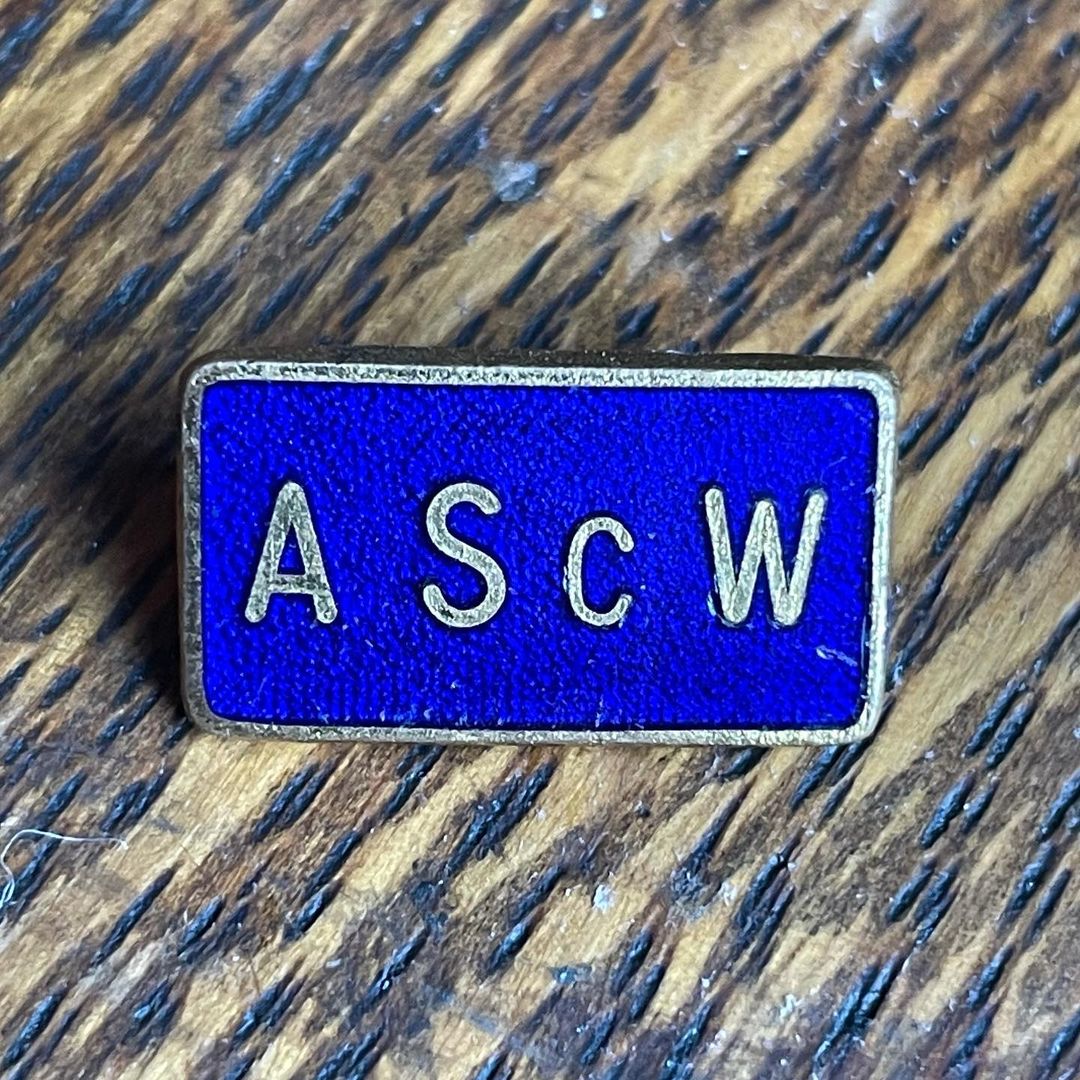 A blue enamel badge with AScW, that stands for Association of Scientific Workers printed on it on a wooden background.